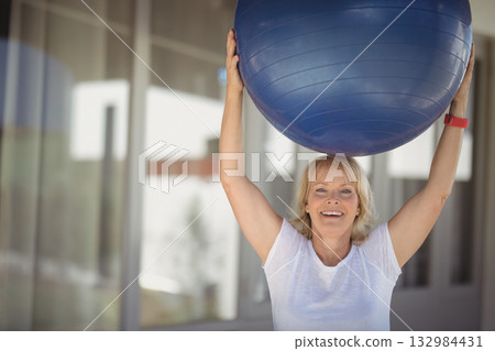 Senior woman lifting blue exercise ball overhead in gym wearing red fitness tracker, copy space Senior woman lifting blue exercise ball overhead in gym wearing red fitness tracker, copy space 132984431