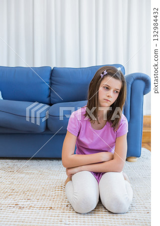 Female child kneeling, gazing down on rug beside blue sofa, wearing purple hair clip, copy space 132984432