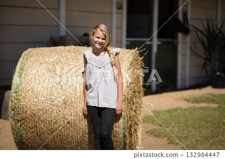Teenage girl standing and leaning against hay bale in front of barn door near potted plant 132984447