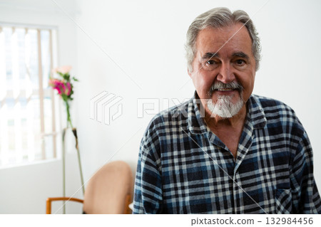 Senior man standing in living room wearing checkered shirt beside vase with flowers, copy space 132984456
