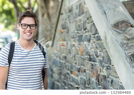 Man standing by stone wall on sidewalk wearing striped tee eyeglasses backpack, copy space 132984460