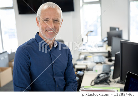 Senior man wearing blue shirt leaning against desk smiling in office with laptop, copy space 132984463
