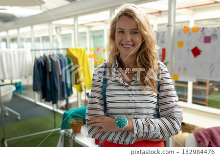 Female fashion designer crossing arms and smiling in studio with tape measure and clothing rack 132984476