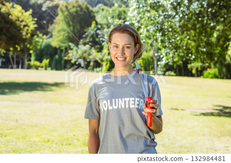Smiling woman standing in open park wearing VOLUNTEER t-shirt and holding red litter picker tool 132984481