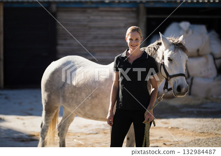 Standing next to white horse in sunlit stable yard holding lead rope in riding clothes 132984487