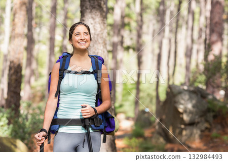 Woman standing and looking upward on sunlit dirt forest trail holding trekking poles and backpack 132984493