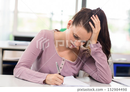 Woman wearing mauve long-sleeve top writing with pen on paper at classroom desk while resting head Woman wearing mauve long-sleeve top writing with pen on paper at classroom desk while resting head 132984498