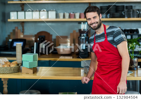 Male barista leaning on wood counter in cafe with bread basket and espresso machine, copy space 132984504
