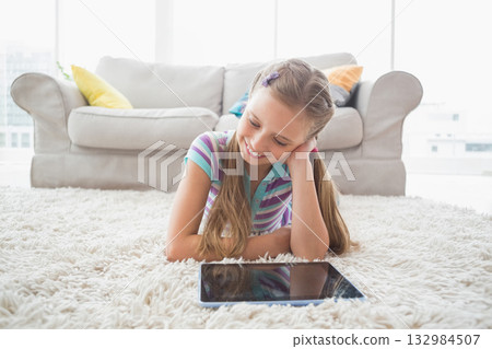 Girl lying on white shag carpet in front of gray sofa with colorful pillows using tablet Girl lying on white shag carpet in front of gray sofa with colorful pillows using tablet 132984507