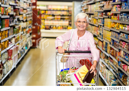 Senior female customer pushing shopping cart down grocery aisle, carrying cereal box and pineapple Senior female customer pushing shopping cart down grocery aisle, carrying cereal box and pineapple 132984515