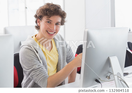 Male student sitting at white desk in computer lab operating silver monitor and typing on keyboard 132984518