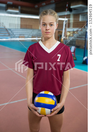 Teenage female standing at volleyball net in gym, holding volleyball by waist in jersey number 7 132984519