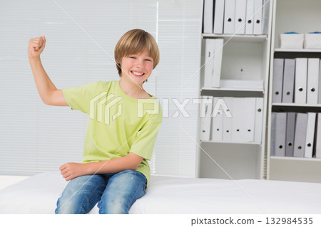 School-age boy flexing right arm while sitting on exam table under window blinds in exam room 132984535
