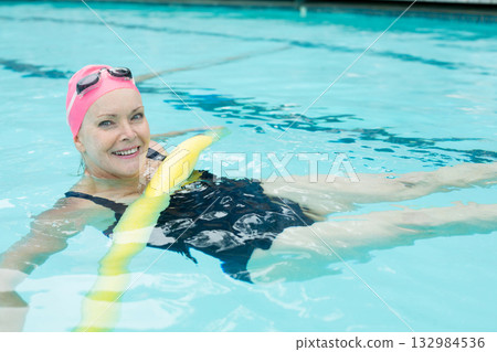 Senior woman floating on yellow noodle at outdoor pool wearing black swimsuit pink swim cap goggles Senior woman floating on yellow noodle at outdoor pool wearing black swimsuit pink swim cap goggles 132984536