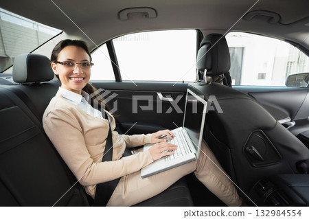 Woman sitting in backseat of modern sedan wearing eyeglasses and seatbelt typing on laptop on lap Woman sitting in backseat of modern sedan wearing eyeglasses and seatbelt typing on laptop on lap 132984554