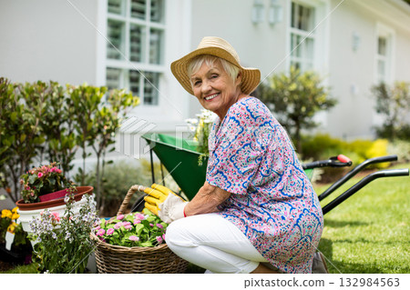 Senior woman kneeling on yard arranging flower pots near wheelbarrow wearing straw hat, copy space 132984563