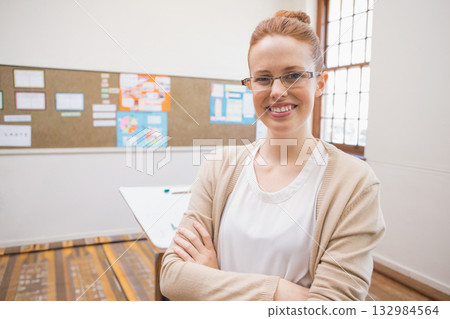 Female instructor standing with arms crossed in classroom by window near bulletin board and papers 132984564