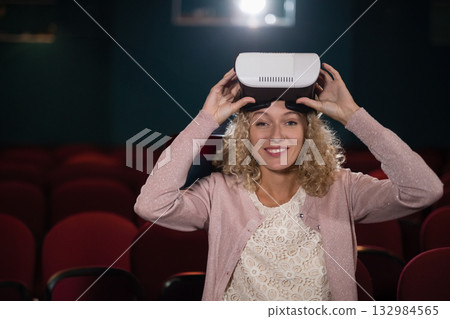 Woman sitting among red auditorium seats positioning virtual reality headset on head at theater Woman sitting among red auditorium seats positioning virtual reality headset on head at theater 132984565