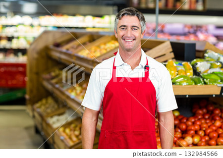 Smiling male grocery store employee wearing red apron standing in produce aisle with green peppers 132984566