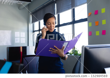 Asian woman office worker holding binder and speaking into headset microphone in open-plan office Asian woman office worker holding binder and speaking into headset microphone in open-plan office 132984569