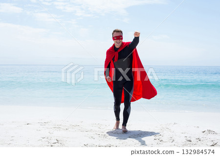 Mid adult man in black bodysuit and red cape and eye-mask standing on beach raising fist 132984574