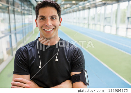Male athlete standing arms crossed on track at sports facility wearing earphones and armband 132984575