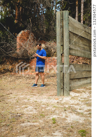 Man wearing blue gear climbing wooden obstacle wall outdoors beside collapsed wall with earphones 132984577