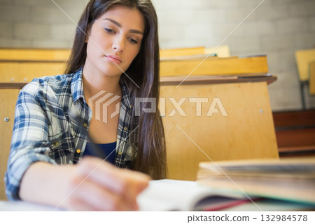 Female student leaning forward while writing on paper at lecture hall desk with books, copy space 132984578