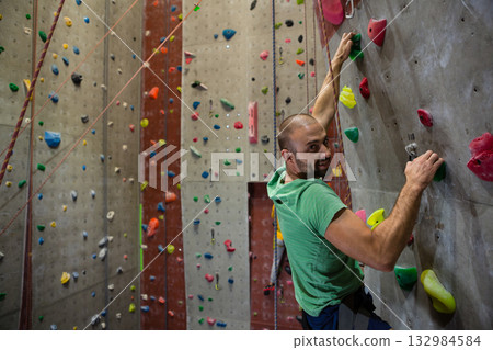 Man climbing artificial rock wall in gym gripping multicolored holds with rope harness, copy space 132984584