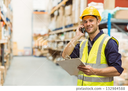 Man in safety vest, hard hat in warehouse aisle using clipboard, calling smartphone, copy space 132984588