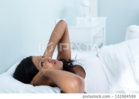Woman reclining on bed, hands behind head, in bedroom near bedside table with pitcher, copy space 132984589