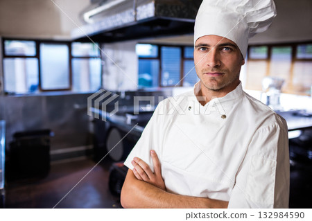 Male chef standing with crossed arms under hood in restaurant kitchen wearing white coat and hat 132984590