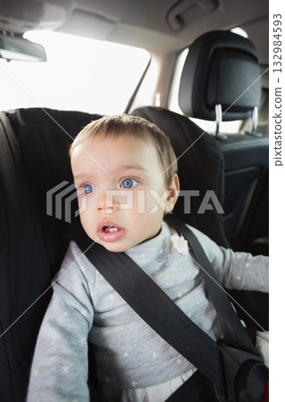 Infant boy sitting in car seat secured by black five-point harness beside back seat car headrest 132984593