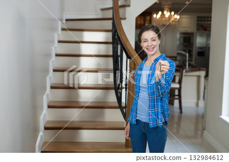 White woman in blue shirt holding keys at foyer with staircase, kitchen island, fridge, copy space 132984612