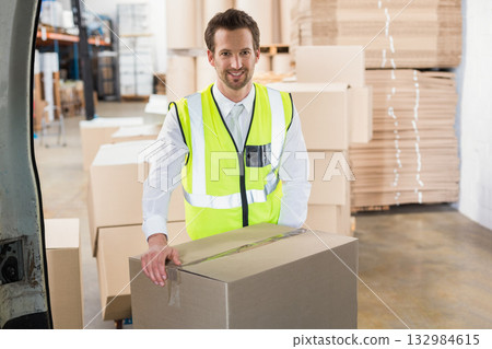 Male worker standing in warehouse behind cardboard box at delivery van rear wearing safety vest 132984615