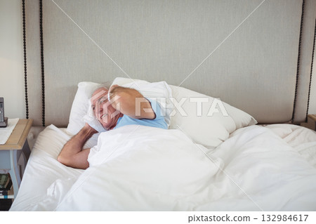 Senior man lying in bedroom pressing pillow over ears beside nightstand with telephone, copy space Senior man lying in bedroom pressing pillow over ears beside nightstand with telephone, copy space 132984617