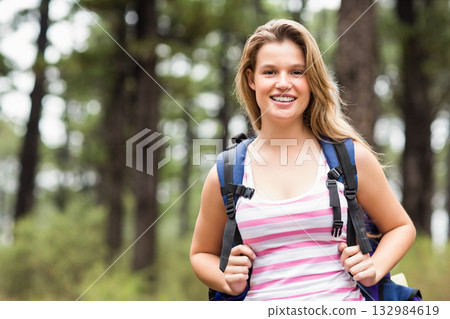 Woman wearing sleeveless pink striped top smiling at camera in pine forest holding hiking backpack 132984619