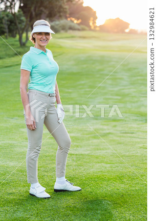 Senior woman standing on golf fairway at sunset wearing visor, golf glove, golf shoes, copy space 132984621