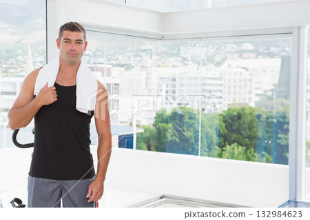 Adult male towel-shouldered leaning by machine handle in gym by windows showing skyline, copy space Adult male towel-shouldered leaning by machine handle in gym by windows showing skyline, copy space 132984623