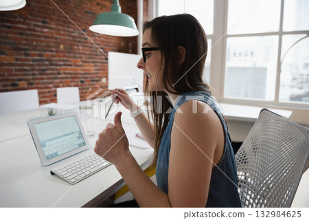 Woman holding stylus above tablet and giving thumbs up at modern office workspace Woman holding stylus above tablet and giving thumbs up at modern office workspace 132984625