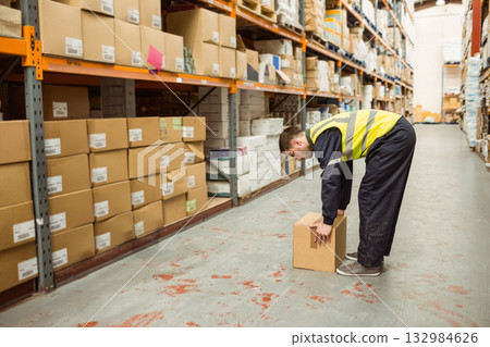 Male worker in high-visibility vest bending and lifting cardboard box on aisle with shelving racks 132984626