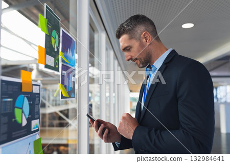 Mature adult man in suit standing next to glass wall tapping smartphone with charts, sticky notes 132984641