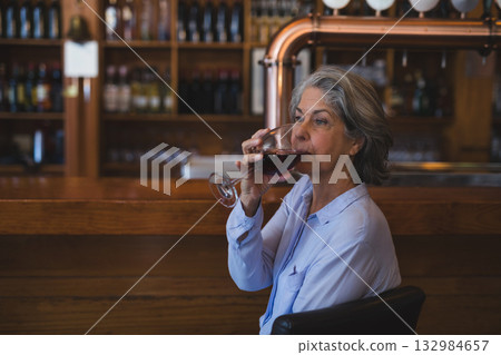 Senior woman raising stemmed wine glass to lips at bar counter with liquor bottles, copy space 132984657