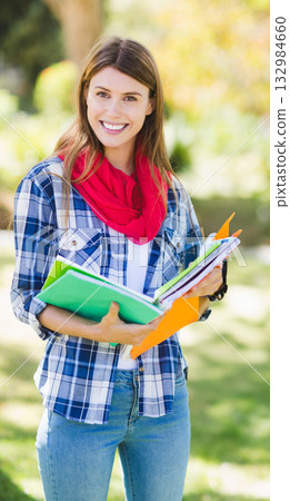 Female student standing on campus lawn holding notebooks and folder showing backpack strap and pen 132984660