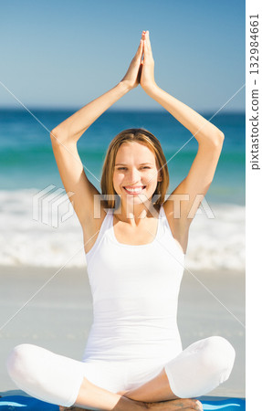 Woman practicing yoga wearing white sportswear sitting cross-legged on blue yoga mat on sandy beach 132984661