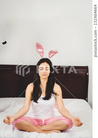 Asian woman sitting cross legged meditating on bed with white sheets wearing pink bunny ears 132984664