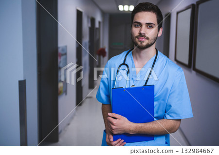Male health worker standing in corridor holding blue clipboard and wearing stethoscope, copy space Male health worker standing in corridor holding blue clipboard and wearing stethoscope, copy space 132984667