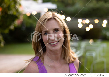 Woman in purple sleeveless top smiling and standing in garden under string lights near round table 132984670