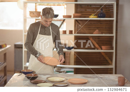 Senior male potter standing at wooden worktable holding ceramic plate in ceramics workshop Senior male potter standing at wooden worktable holding ceramic plate in ceramics workshop 132984671