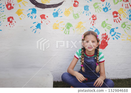 Child girl sitting cross-legged on grass in front of handprint wall holding red paint, copy space 132984688